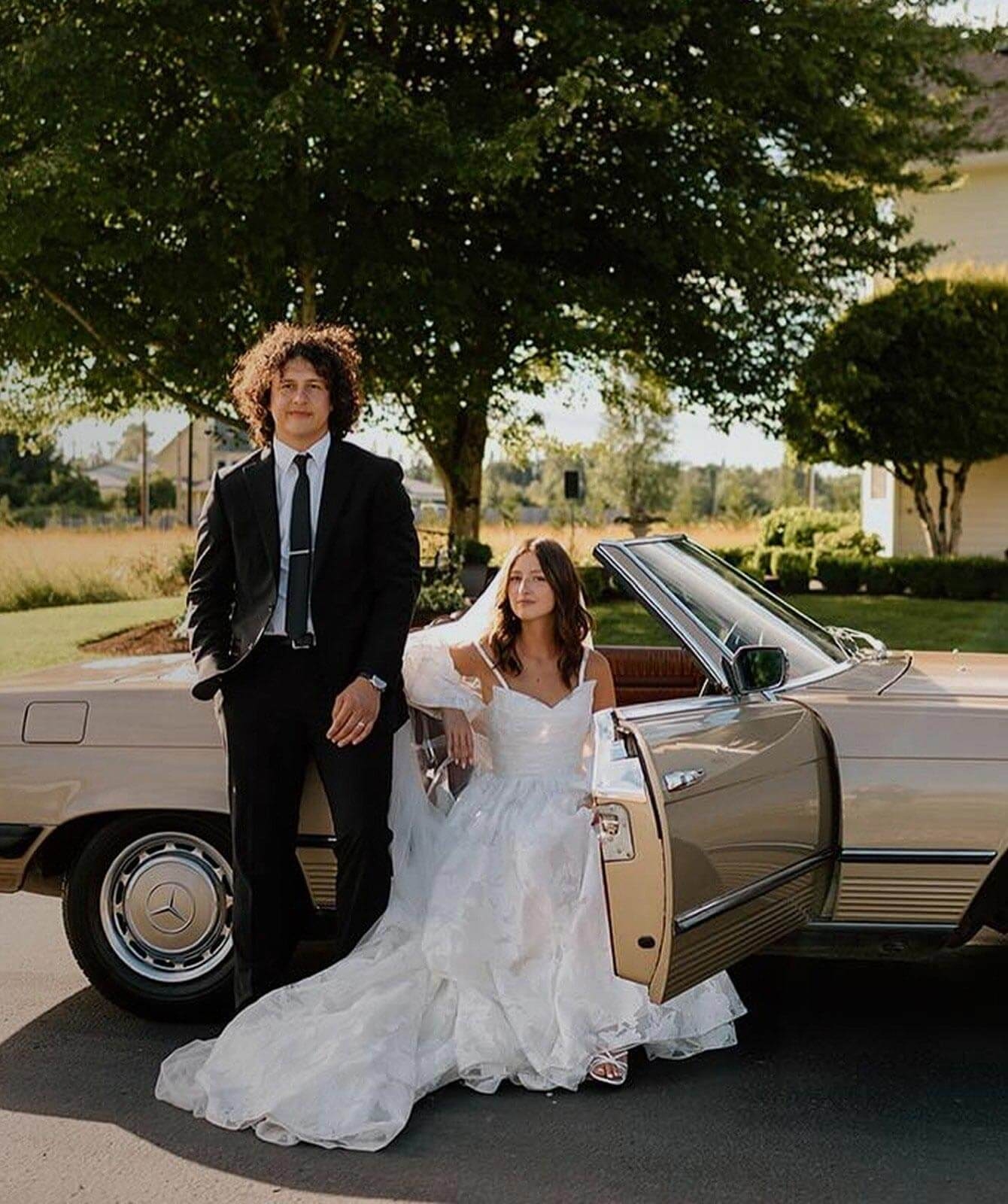 Сouple wearing a white gown and a black suit near a car. Mobile image